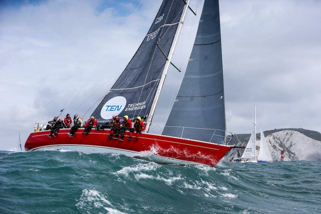 The red hull of Scarlet Oyster surfing a wave while sailing upwind with land and another yacht in the background.