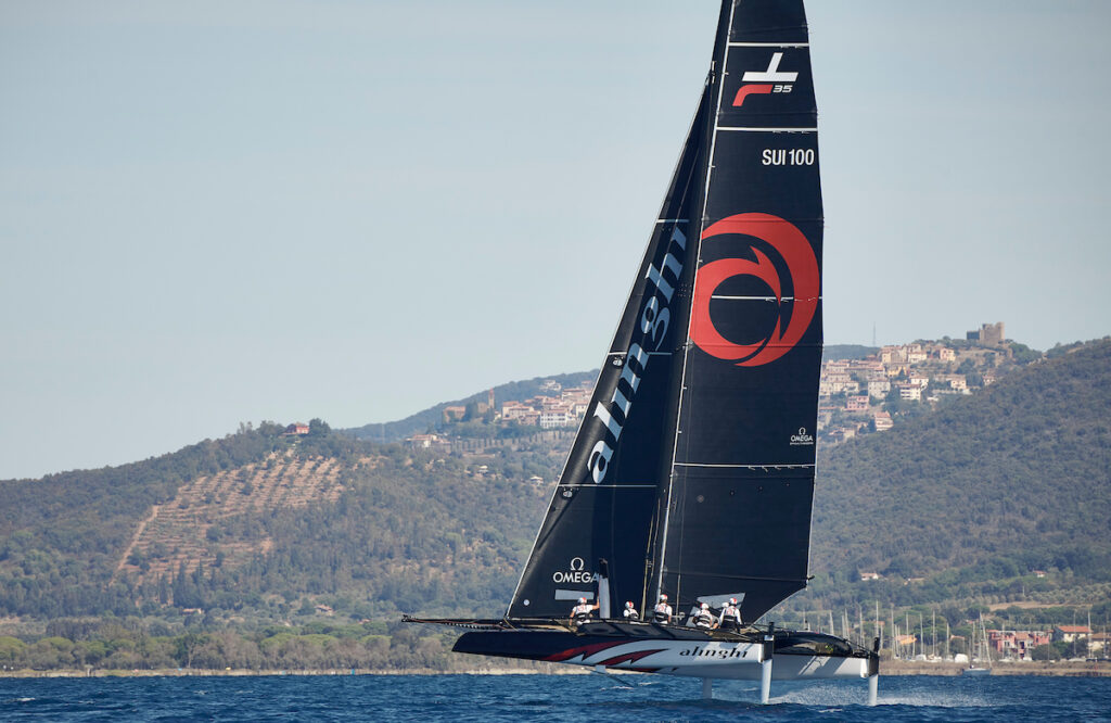 Alinghi sailing upwind, mountains and buildings in the background.