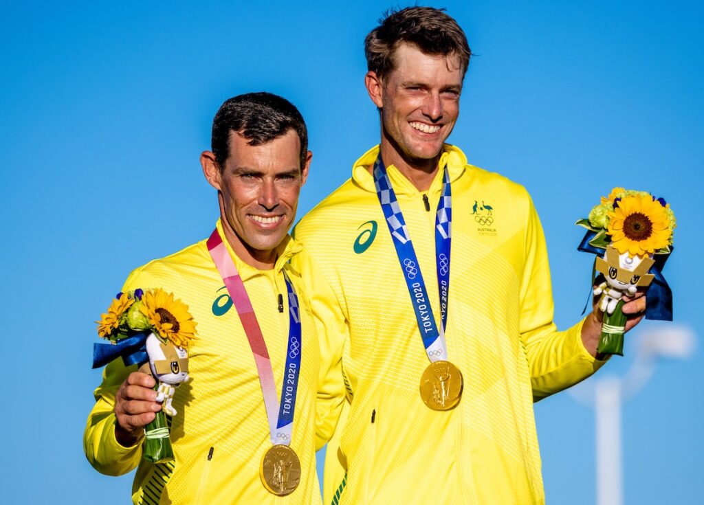 Mat Belcher and Will Ryan smiling at the medal ceremony with their medals around their necks and holding flowers.