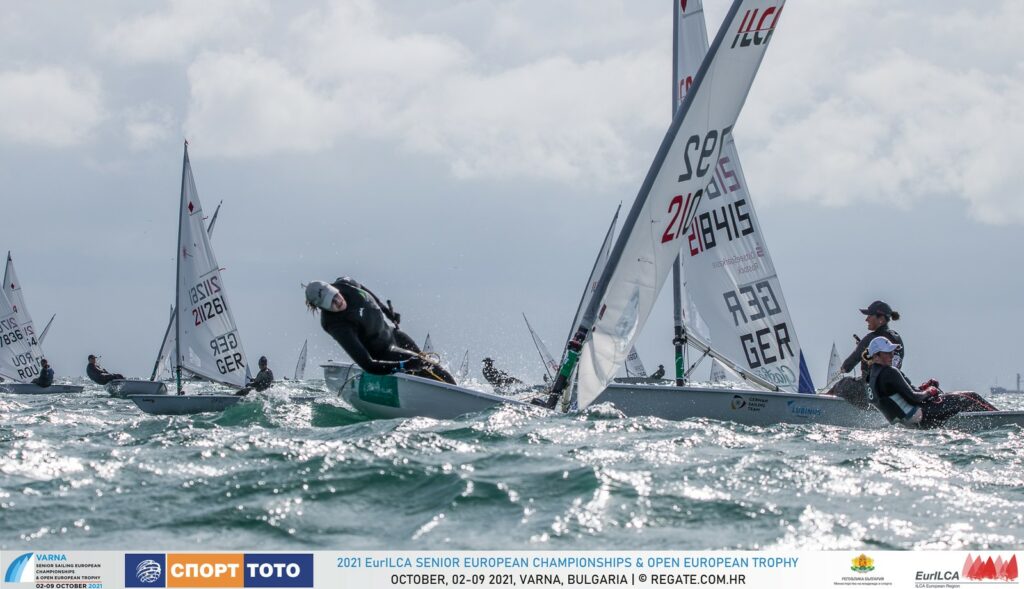 A sailor hiking out just as they finish a tack. The fleet of different tacks behind them.