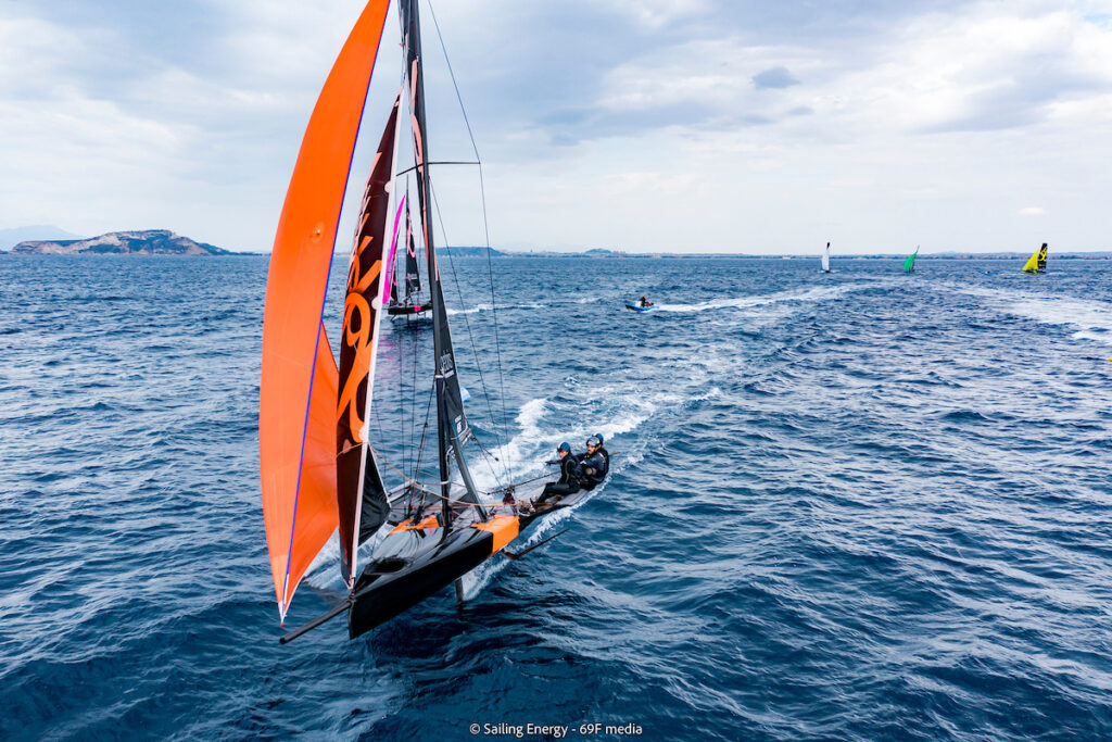 Aerial shot of boat with orange kite on spinnaker reach with fleet behind them (spread out).