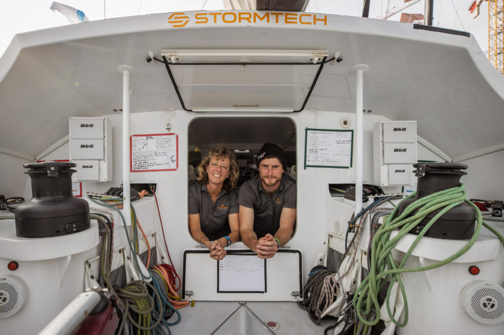 Sam Davies and Nicolas Lunven looking out onto the cockpit from the cabin, smiling.