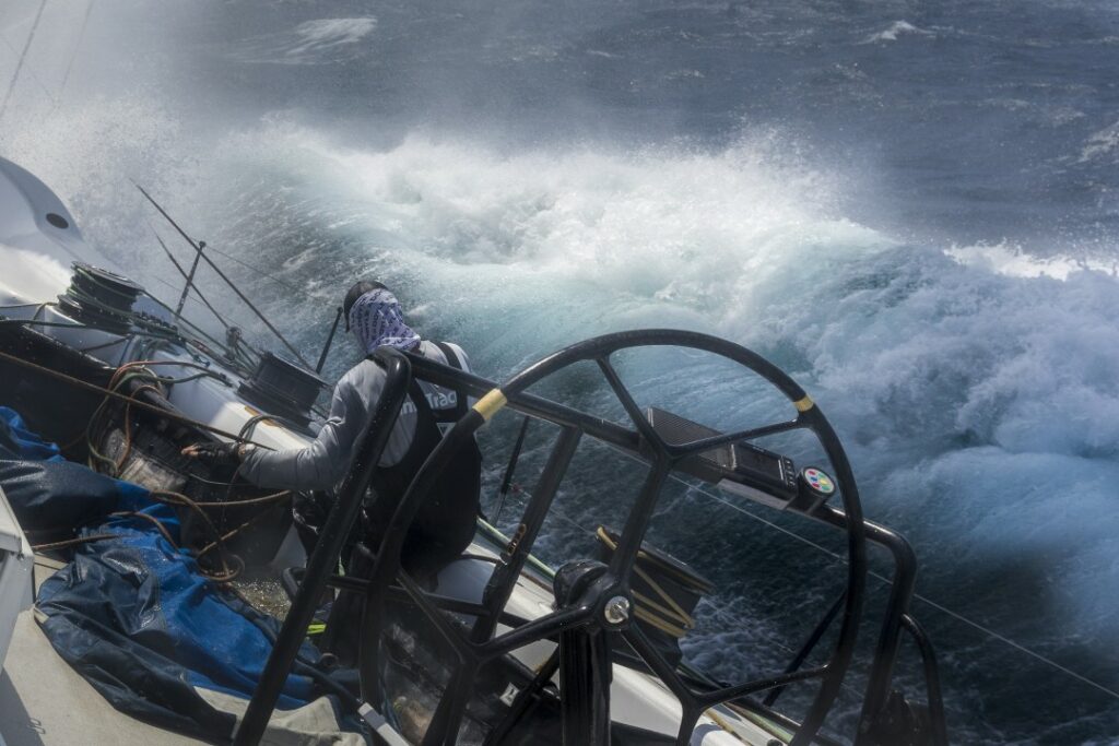 A wave breaking towards the leeward side of the boat where a sailor sits.