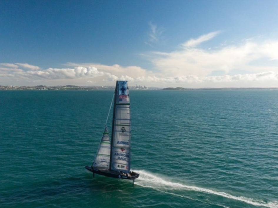 Aerial shot of American Magic foiling upwind on a crystal clear day, with a few puffy clouds in the background.