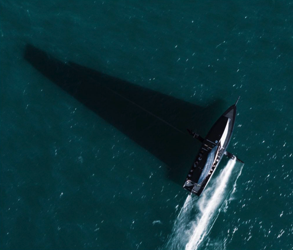 Aerial shot of American magic on the foils. It's sail creates a huge shadow on the water.