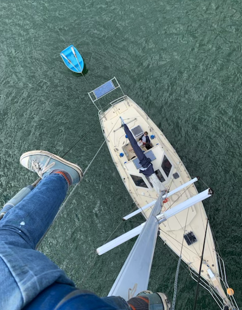 View from the top of the mast looking down at the moored boat, person sitting in cockpit and dinghy out the back.