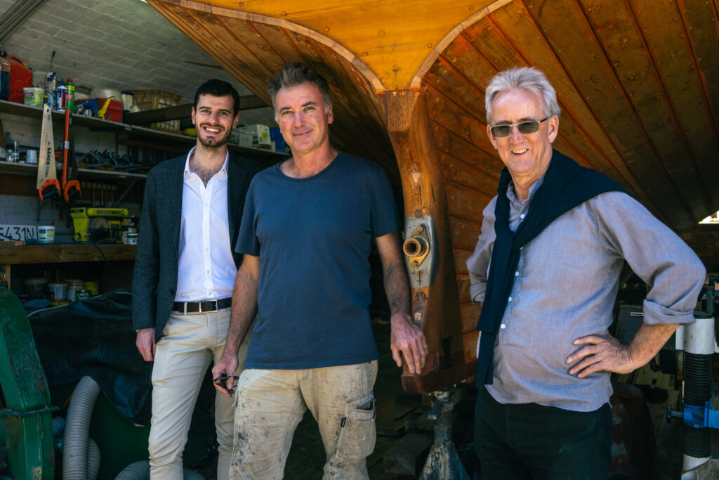 Nathan Cox, Rob Gordon and Jim Atkins standing with a wooden boat in shed behind them.