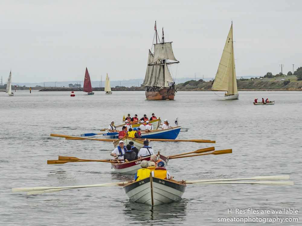 Five rowboats in a line with old sailing yachts in the background.