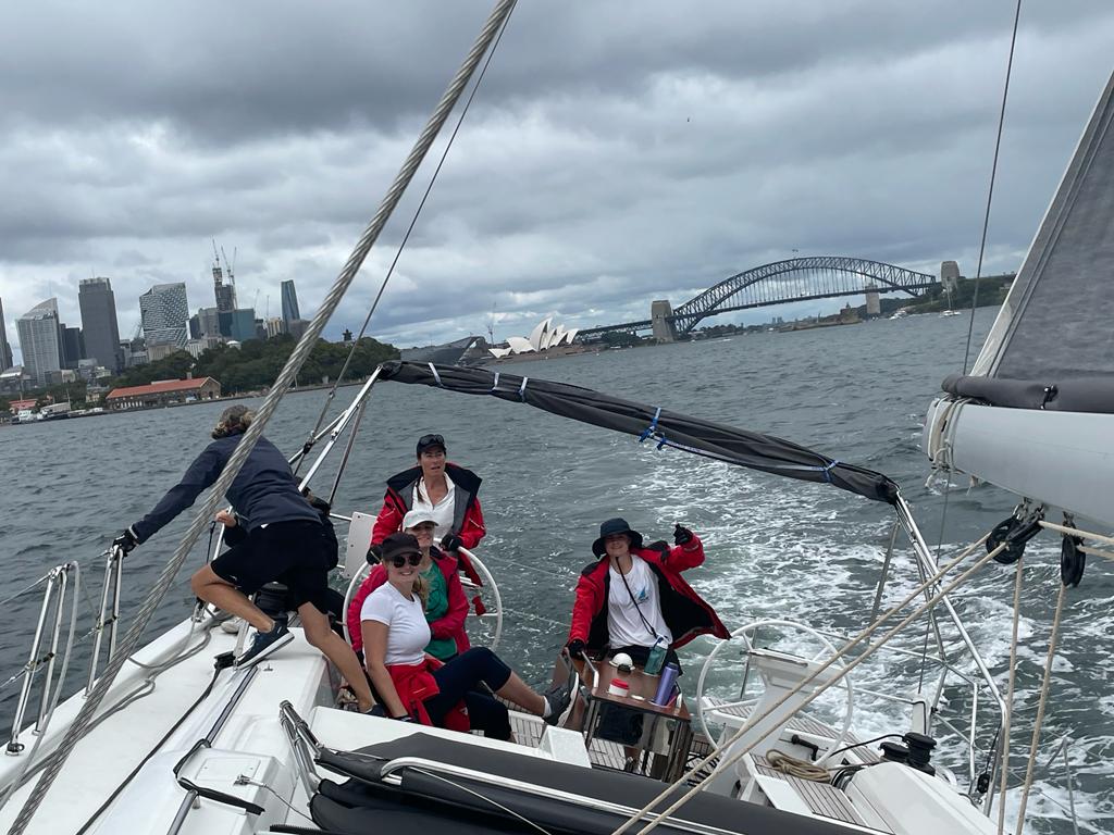 People smiling at the back of a sailing yacht with harbour bridge and opera house in the background.