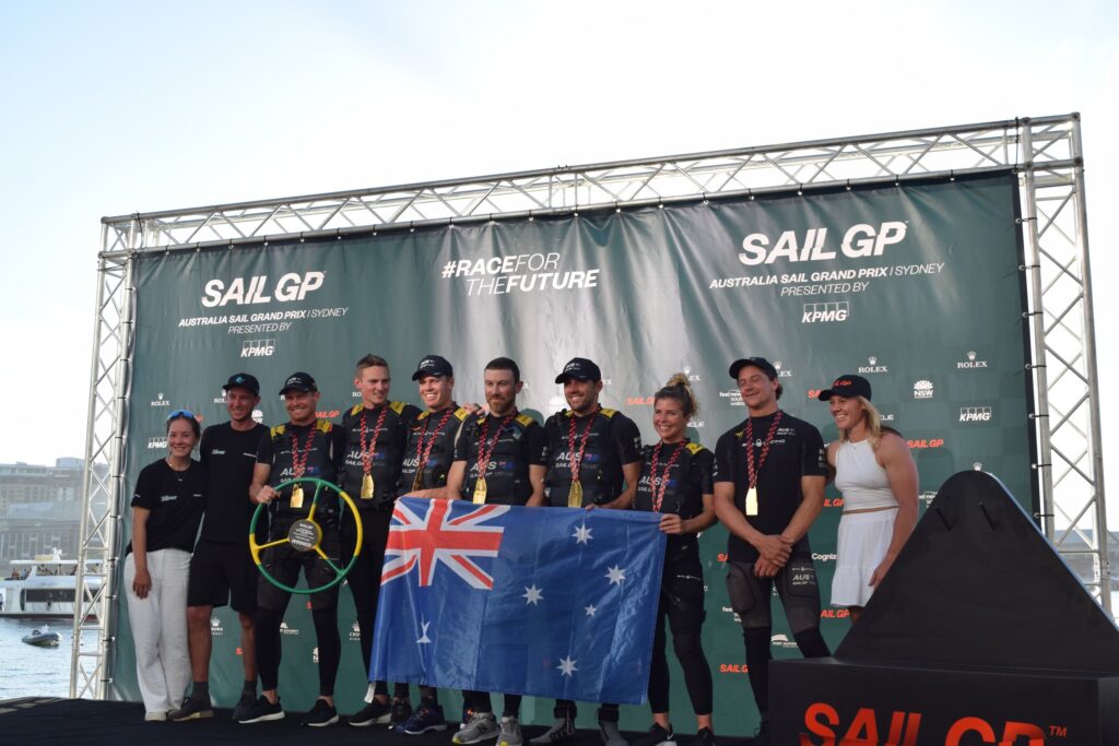 SailGP team holding trophy and flag on stage.