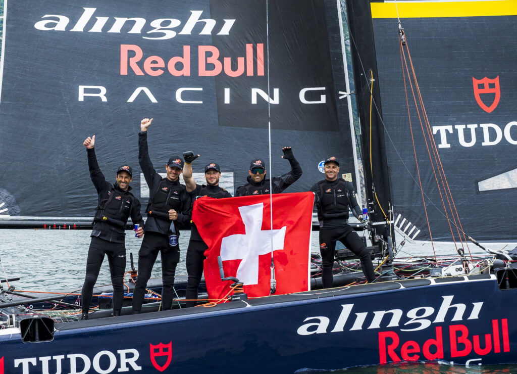 Arnaud Psarofaghis and the winning crew of Alinghi Red Bull Racing - SUI 8 holding flag on boat on water.